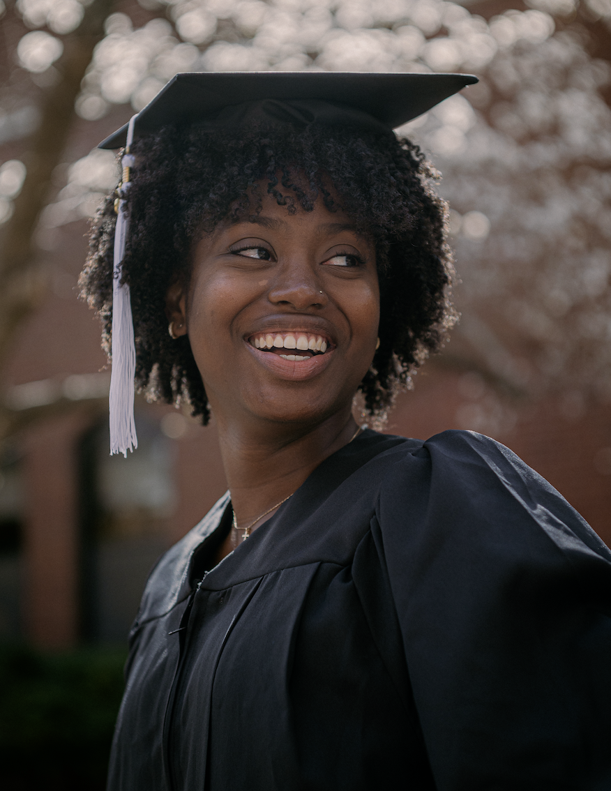 Portrait of a smiling graduating student wearing a black cap and gown, standing outdoors in front of a blooming tree and brick building. The graduate, with curly hair and a white tassel on the cap, looks off to the side, radiating joy and pride.
